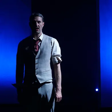 Ein Schauspieler steht halb im Dunkeln auf der Bühne, nur von einem kalten blauen Licht seitlich beleuchtet. Er trägt einen grauen Anzug mit Weste und roter Krawatte, wirkt ernst und konzentriert – eine dramatische, spannungsgeladene Szene.