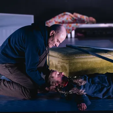 Ein Mann kniet neben einer Frau am Boden, die dramatisch daliegt. Im Hintergrund ist eine weitere Figur auf einem Sofa zu sehen.