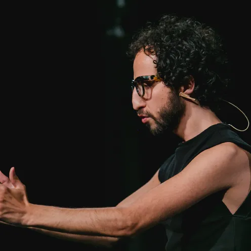 Ein Mann mit Locken und Brille spricht auf einer Bühne, trägt ein Headset-Mikrofon und gestikuliert mit den Händen. Er wirkt konzentriert und leidenschaftlich, die Szene ist dunkel mit Fokus auf seine Person.