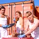 Drei Personen in weißen Schürzen und blauen Handschuhen stehen vor orangenen Containern. Eine Frau trinkt Wein, der Mann hält ein Tablett mit Pinsel, eine andere Frau arbeitet konzentriert an einem Kochtopf auf einem Tisch.