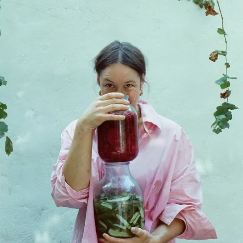 Eine Frau in rosa Hemd hält zwei Gläser mit eingelegtem Gemüse und schaut direkt in die Kamera. Sie steht vor einer hellen Wand mit herabhängenden Weinreben und wirkt ruhig und konzentriert.