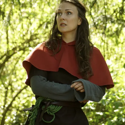 Eine Frau in mittelalterlicher Gewandung steht im sonnendurchfluteten Wald. Sie trägt ein rotes Cape, einen grünen Gürtel und blickt nachdenklich zur Seite. Ihr langer, dunkler Zopf fällt über die Schulter.