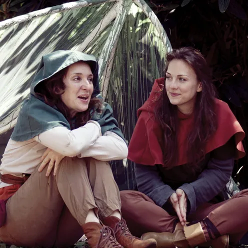 Zwei Frauen in mittelalterlicher Kleidung sitzen entspannt vor einem Zelt und unterhalten sich. Die eine trägt einen grünen Umhang, die andere einen roten. Beide wirken fröhlich und verbunden, umgeben von Natur und Schatten.