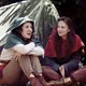 Zwei Frauen in mittelalterlicher Kleidung sitzen entspannt vor einem Zelt und unterhalten sich. Die eine trägt einen grünen Umhang, die andere einen roten. Beide wirken fröhlich und verbunden, umgeben von Natur und Schatten.