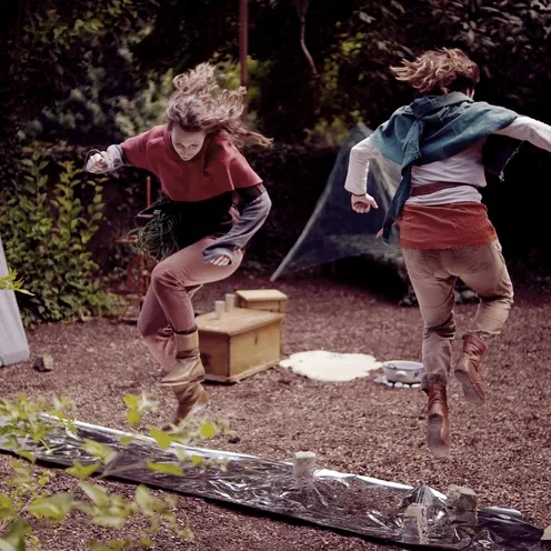 Zwei Frauen in mittelalterlicher Gewandung springen energiegeladen über eine mit Folie bedeckte Strecke. Die Szene spielt in einem Waldlager mit Zelten, Requisiten und einem Banner.