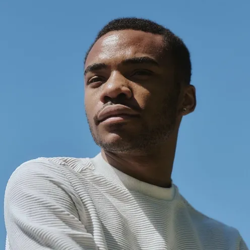 Ein Mann blickt nachdenklich in die Ferne. Er trägt einen weißen Pullover mit Strukturmuster. Der Himmel im Hintergrund ist klar und blau, die Sonne wirft sanfte Schatten auf sein Gesicht. Ein dezenter Ohrstecker rundet den Look ab.