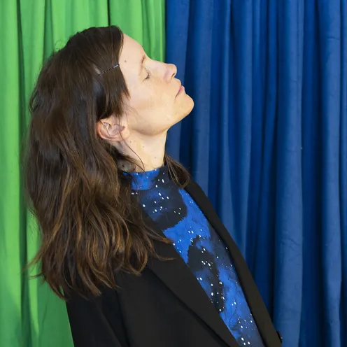 Eine Frau mit geschlossenen Augen und leicht nach hinten geneigtem Kopf steht seitlich vor einem Hintergrund aus grünen und blauen Vorhängen. Sie trägt ein schwarzes Sakko und ein blau gemustertes Oberteil.