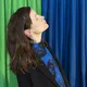 Eine Frau mit geschlossenen Augen und leicht nach hinten geneigtem Kopf steht seitlich vor einem Hintergrund aus grünen und blauen Vorhängen. Sie trägt ein schwarzes Sakko und ein blau gemustertes Oberteil.