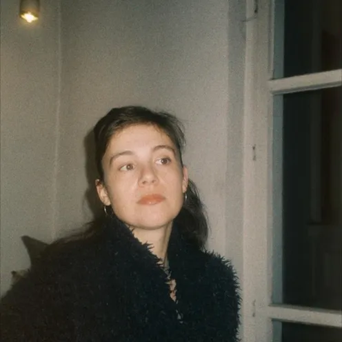 Eine Frau mit dunklem, zurückgekämmtem Haar blickt nachdenklich zur Seite. Sie trägt einen flauschigen, dunklen Mantel und steht in einem spärlich beleuchteten Raum neben einem Fenster. Die Atmosphäre wirkt ruhig und intim.