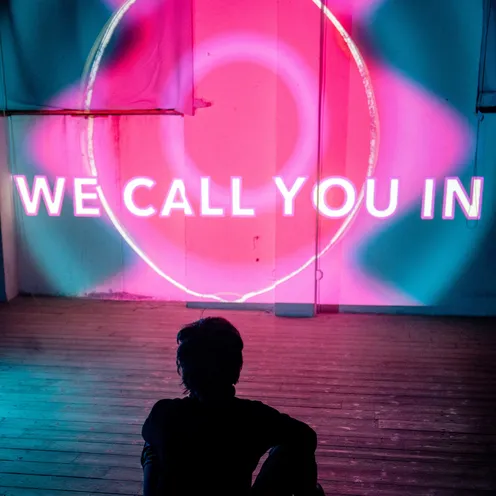 Eine Person sitzt auf Holzboden vor einer Wand mit leuchtender Projektion in Pink und Blau. Die Worte „WE CALL YOU IN“ erscheinen groß in der Mitte des Bildes, eingerahmt von einem kreisförmigen Lichtmuster.