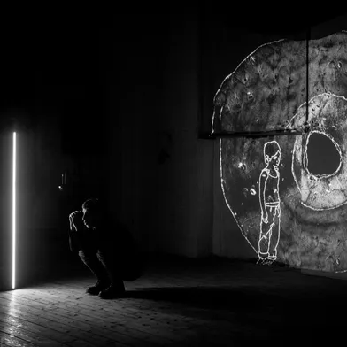 In einem dunklen Raum hockt eine Person vor einem leuchtenden Lichtstreifen. An der Wand ist eine Projektion mit einem gezeichneten Kind und einer strukturierten Fläche zu sehen – die Szene wirkt künstlerisch und nachdenklich.