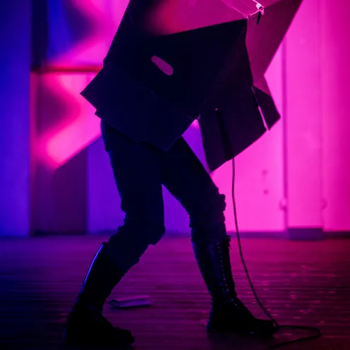 Eine Person steht in dramatischer Pose auf einer Bühne mit pink-violetter Beleuchtung. Sie trägt schwarze Stiefel und hat einen großen schwarzen Karton über dem Oberkörper – möglicherweise Teil einer Performance oder Kunstaktion.