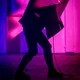 Eine Person steht in dramatischer Pose auf einer Bühne mit pink-violetter Beleuchtung. Sie trägt schwarze Stiefel und hat einen großen schwarzen Karton über dem Oberkörper – möglicherweise Teil einer Performance oder Kunstaktion.