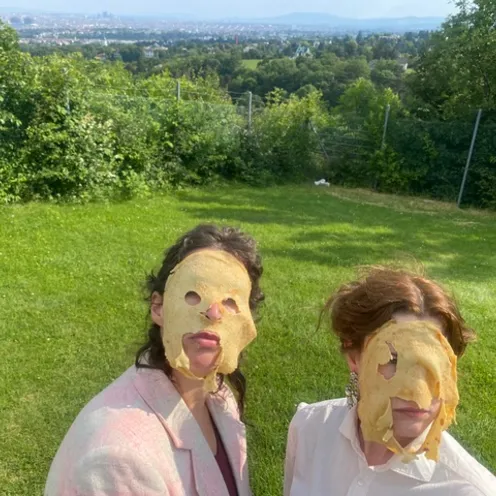 Zwei Personen stehen auf einer Wiese mit Blick auf eine Stadtlandschaft im Hintergrund. Sie tragen Masken aus flachen Broten mit ausgeschnittenen Löchern. Die Szene wirkt skurril und humorvoll inszeniert.