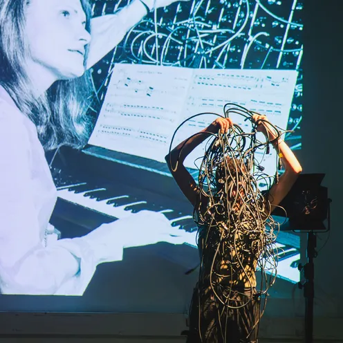 Eine Person steht vor einer Projektion mit einem Bild einer Frau am Synthesizer. Sie hält ein großes Knäuel aus Kabeln in die Luft, wodurch ihr Gesicht verdeckt ist. Die Szene wirkt kreativ, experimentell und technisch.