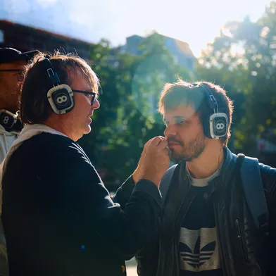 Zwei Männer mit Kopfhörern stehen sich gegenüber, einer berührt fürsorglich das Gesicht des anderen. Sonnenlicht fällt durch Bäume im Hintergrund, die Szene wirkt intim und konzentriert.