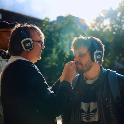 Zwei Männer mit Kopfhörern stehen sich gegenüber, einer berührt fürsorglich das Gesicht des anderen. Sonnenlicht fällt durch Bäume im Hintergrund, die Szene wirkt intim und konzentriert.