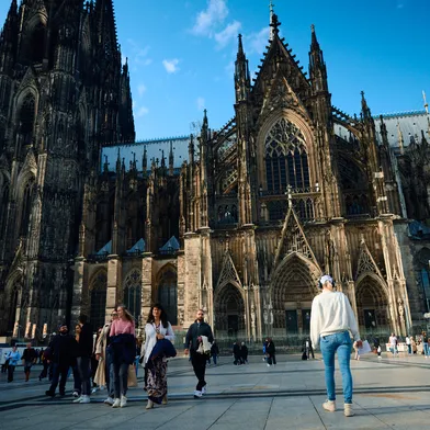 Vor dem imposanten Kölner Dom spazieren Menschen über den sonnigen Vorplatz. Die gotische Fassade ragt majestätisch in den blauen Himmel, während Passanten die historische Atmosphäre genießen.