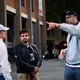 Bildbeschreibung sagte: Drei Personen stehen in einer Stadtumgebung und unterhalten sich ernst. Einer zeigt in eine Richtung, während der Mann in der Mitte mit Kopfhörern in der Hand nachdenklich schaut. Die Stimmung wirkt konzentriert und leicht angespannt.