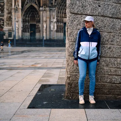 Eine Frau in sportlicher Kleidung lehnt an einem massiven Steinsockel auf einem leeren Platz. Im Hintergrund ist unscharf der Kölner Dom zu erkennen. Die Szene wirkt ruhig und nachdenklich.
