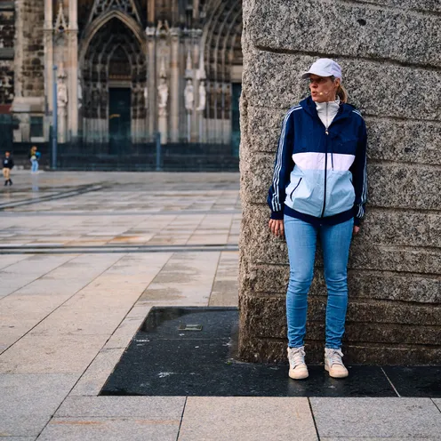 Eine Frau in sportlicher Kleidung lehnt an einem massiven Steinsockel auf einem leeren Platz. Im Hintergrund ist unscharf der Kölner Dom zu erkennen. Die Szene wirkt ruhig und nachdenklich.
