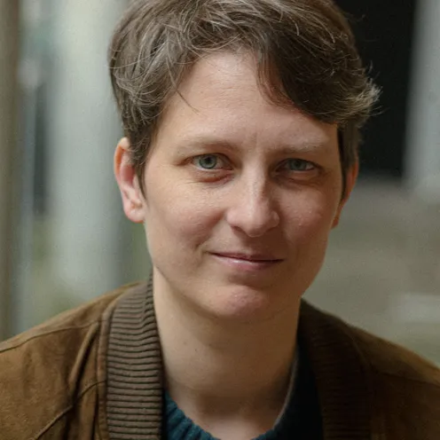 Eine Person mit kurzem, braunem Haar blickt freundlich in die Kamera. Sie trägt eine braune Jacke über einem dunkelgrünen Pullover. Der Hintergrund ist unscharf, wodurch das Gesicht im Fokus steht. Die Lichtstimmung ist natürlich und weich.