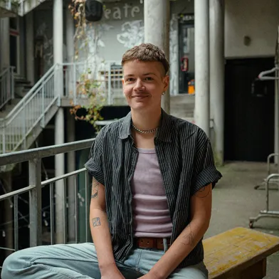 Eine Person mit kurzem Haar sitzt lässig auf einer Bank in einem urbanen Innenhof. Sie trägt ein gestreiftes Hemd, ein rosafarbenes Top und helle Jeans. Sichtbare Tattoos und Schmuck unterstreichen den individuellen Stil.