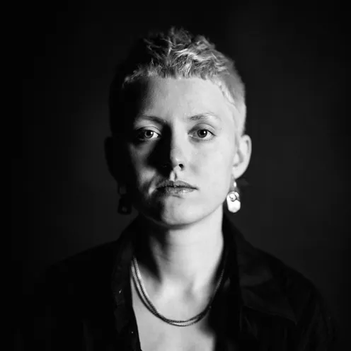 Schwarz-weiß-Porträt einer Person mit kurzem, hellem Haar. Der Blick ist ernst, der Hintergrund dunkel. Sie trägt auffällige Ohrringe, eine Halskette, ein offenes Hemd und ein Top. Das Licht betont die Gesichtszüge dramatisch.