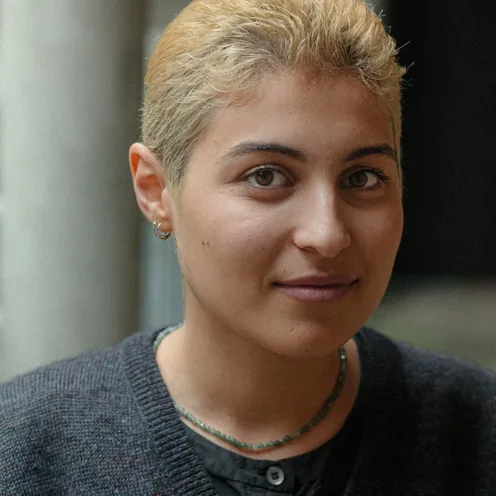 Eine Person mit kurz blond gefärbtem Haar blickt ruhig in die Kamera. Sie trägt einen dunklen Pullover, ein schwarzes Hemd und eine grüne Perlenkette. Der Hintergrund ist unscharf, wodurch das Gesicht im Mittelpunkt steht.