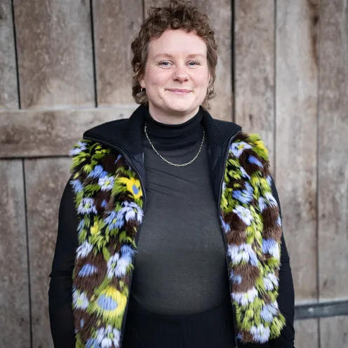 Bildbeschreibung sagte: Eine Person mit lockigem, kurzem Haar steht vor einer Holzwand und lächelt. Sie trägt ein schwarzes Oberteil mit transparenter Struktur und eine bunte Kunstfellweste in Grün, Braun, Lila und Weiß – ein ausdrucksstarker Look.