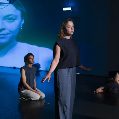 Drei Performer:innen in nachdenklicher Pose auf einer dunklen Bühne, im Hintergrund eine große Projektion eines Gesichts in Blau. Die Szene wirkt intensiv und künstlerisch inszeniert, mit starker Licht- und Bildwirkung.