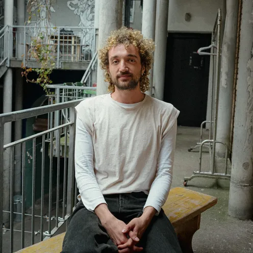 Eine Person mit lockigem, blondem Haar sitzt auf einer Holzbank in einem urbanen Innenhof. Sie trägt ein weißes Langarmshirt und schwarze Jeans. Der Blick ist ruhig, die Hände sind gefaltet, der Hintergrund zeigt ein kreativ gestaltetes Gebäude.