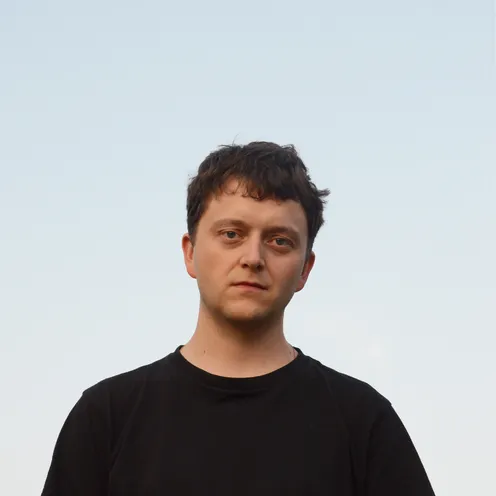 Bildbeschreibung sagte: Porträt eines jungen Mannes mit kurzem, braunem Haar und neutralem Gesichtsausdruck. Er trägt ein schwarzes T-Shirt und steht vor klarem, hellblauem Himmel. Die Stimmung wirkt ruhig und nachdenklich.