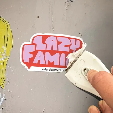 Ein Aufkleber mit der Aufschrift „Lazy Family – oder das Recht auf...“ wird mit einem Schaber von einer grauen Wand entfernt. Links ist ein Teil einer gelben Zeichnung zu sehen. Die Szene wirkt urban und protestartig.