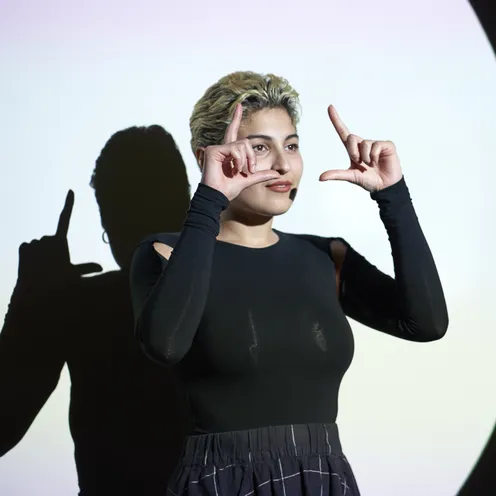 Eine Person mit blondiertem Kurzhaarschnitt steht vor einer Wand mit Lichtprojektion und formt mit den Händen ein Rechteck. Sie trägt ein schwarzes Oberteil mit Cut-outs und wirft einen deutlichen Schatten auf die Wand.