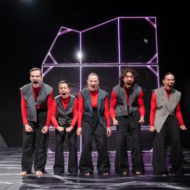 Bildbeschreibung sagte: Fünf barfüßige Performer schreien ausdrucksstark auf einer Bühne mit futuristischem Lichtobjekt im Hintergrund. Sie tragen rote Shirts mit grauen Westen und wirken intensiv und synchron in ihrer Körpersprache.