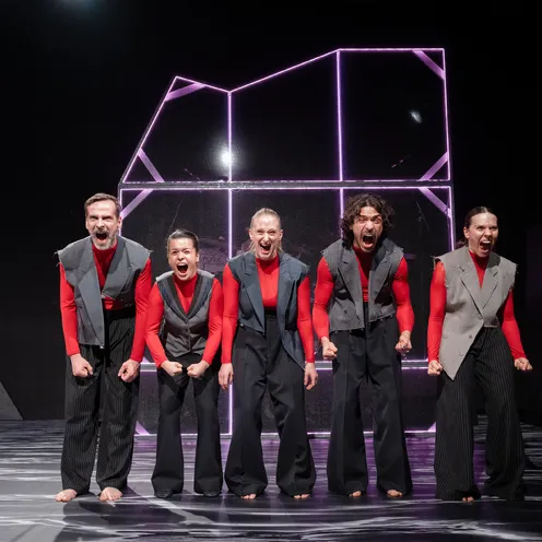 Bildbeschreibung sagte: Fünf barfüßige Performer schreien ausdrucksstark auf einer Bühne mit futuristischem Lichtobjekt im Hintergrund. Sie tragen rote Shirts mit grauen Westen und wirken intensiv und synchron in ihrer Körpersprache.
