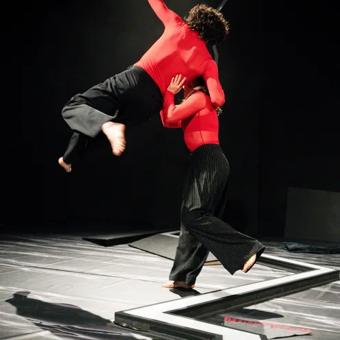 Bildbeschreibung sagte: Zwei Tänzer in roten Oberteilen und dunklen Hosen führen eine dynamische Hebefigur auf. Eine Person hebt die andere kraftvoll in die Luft, der Moment wirkt energiegeladen und spannungsvoll auf der minimalistisch gestalteten Bühne.