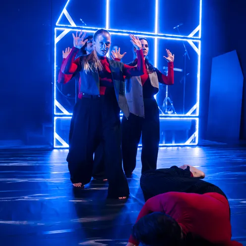 Drei Performer stehen mit erhobenen Händen vor einer leuchtenden, blauen Lichtstruktur, während eine Person am Boden liegt. Die Szene wirkt dramatisch und spannungsvoll, geprägt von intensiver Mimik und kontrastreicher Beleuchtung.