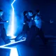 Vier Performer bewegen sich dynamisch durch blaues Licht und Nebel auf einer futuristisch beleuchteten Bühne. Die Szene wirkt geheimnisvoll und intensiv, mit starken Kontrasten und fließender Gruppenchoreografie.