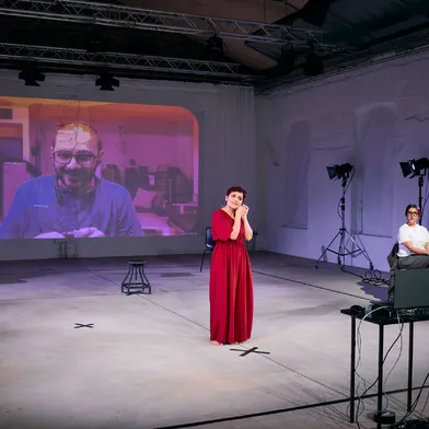 Eine Frau im roten Kleid steht barfuß auf einer Bühne und spricht emotional, im Hintergrund ist ein Mann per Videoprojektion zugeschaltet. Eine weitere Frau sitzt seitlich und beobachtet die Szene.