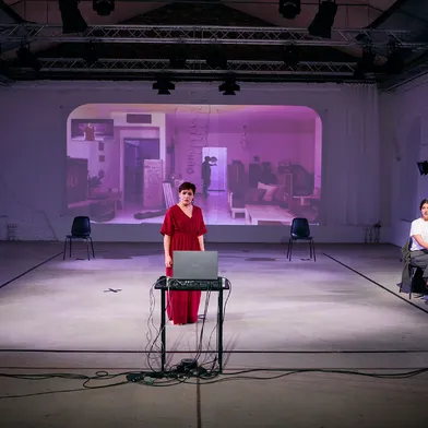 Die Frau im roten Kleid steht ernst vor einem Laptop, während eine Videoprojektion im Hintergrund ein Wohnzimmer zeigt. Die zweite Frau sitzt rechts auf einem Stuhl, mehrere Scheinwerfer sind sichtbar.