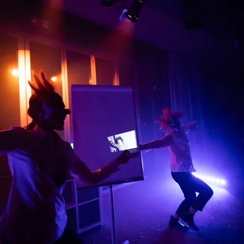 Zwei Personen mit gruseligen Clownmasken tanzen in bunter Bühnenbeleuchtung. Eine projiziert ein Bild auf ein Flipchart, während die andere in dramatischer Pose im Licht steht. Nebel und Farben erzeugen eine düstere Atmosphäre.
