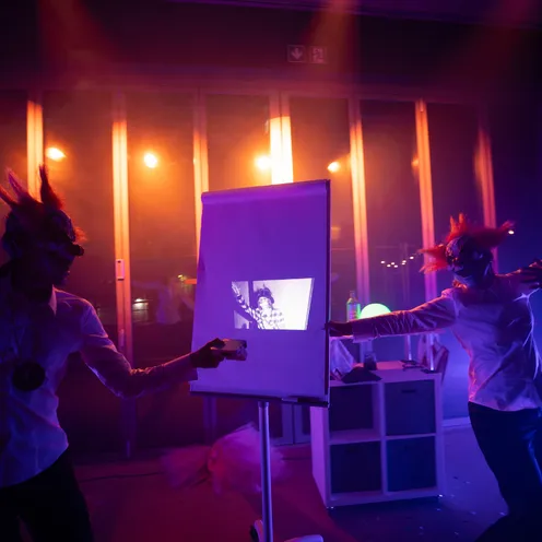 Zwei Personen mit Clownmasken tanzen vor einer Flipchart-Leinwand, auf die ein s/w-Bild projiziert wird. Farbige Bühnenlichter und Nebel schaffen eine geheimnisvolle, theatralische Atmosphäre.