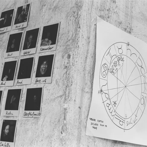An einer Wand kleben Polaroid-Fotos mit handschriftlichen Namen und Begriffen sowie ein astrologisches Horoskop mit dem Namen „Franz Kafka“ und Geburtsdaten. Die Szene wirkt kreativ und persönlich dokumentiert.