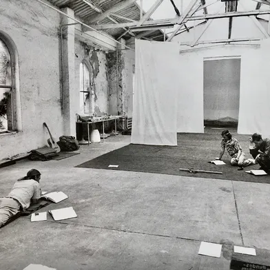 In einer alten Industriehalle mit hohen Decken und abblätternder Wandfarbe sitzen drei Personen auf dem Boden, vertieft in Texte. Bücher und Papiere liegen verstreut, eine entspannte, kreative Atmosphäre dominiert die Szene.