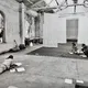 In einer alten Industriehalle mit hohen Decken und abblätternder Wandfarbe sitzen drei Personen auf dem Boden, vertieft in Texte. Bücher und Papiere liegen verstreut, eine entspannte, kreative Atmosphäre dominiert die Szene.