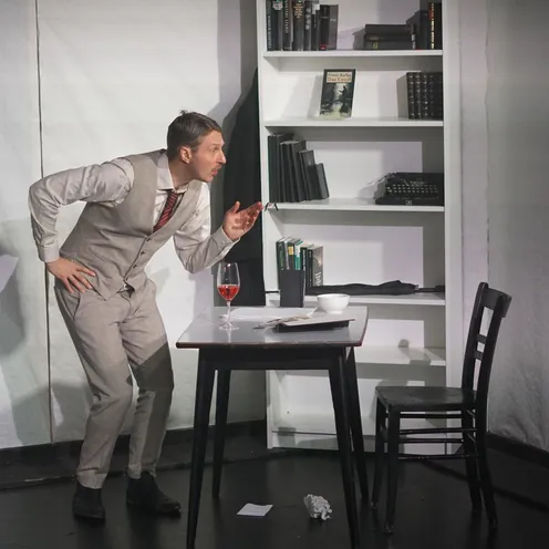 Der Schauspieler beugt sich in Richtung eines Tisches mit Weinglas und Buch, gestikulierend, als führe er ein intensives Gespräch. Im Hintergrund ein Bücherregal mit Requisiten wie Büchern und Schreibmaschine – die Szene wirkt intellektuell und dynamisch.