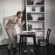 Der Schauspieler beugt sich in Richtung eines Tisches mit Weinglas und Buch, gestikulierend, als führe er ein intensives Gespräch. Im Hintergrund ein Bücherregal mit Requisiten wie Büchern und Schreibmaschine – die Szene wirkt intellektuell und dynamisch.