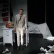 Der Schauspieler steht inmitten einer unordentlichen Bühne: zerknülltes Papier, geöffnete Bücher, ein Koffer. Er lehnt sich an einen Schreibtisch, wirkt erschöpft oder nachdenklich – ein Moment des Stillstands oder der Reflexion.